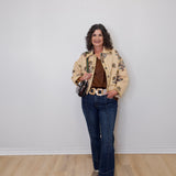Woman wearing a floral jacket, brown top, and blue jeans against a white wall.