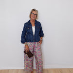 Woman wearing a denim jacket and patterned pants against a white wall.