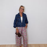 Woman wearing a denim jacket and patterned pants against a white wall.