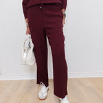 Person wearing a burgundy outfit with white sneakers and a white handbag on a light wooden floor.