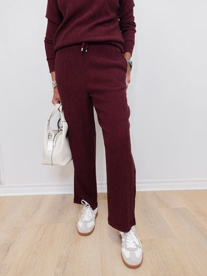 Person wearing a burgundy outfit with white sneakers and a white handbag on a light wooden floor.