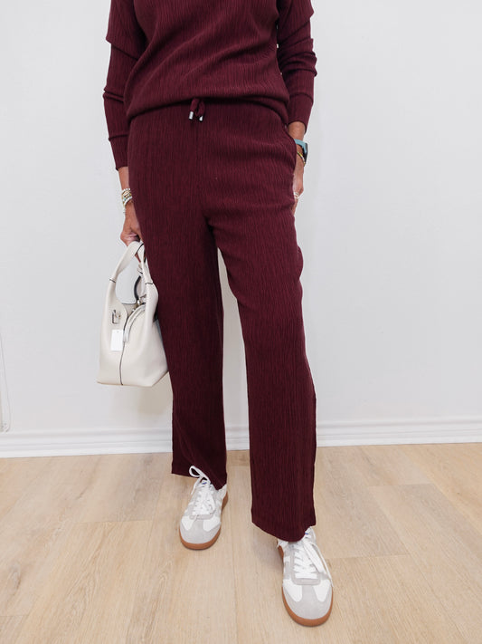 Person wearing a burgundy outfit with white sneakers and a white handbag on a light wooden floor.
