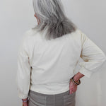 An older woman with long gray hair is shown from behind, wearing a cream-colored long-sleeve top and gray pants, standing against a plain light background.