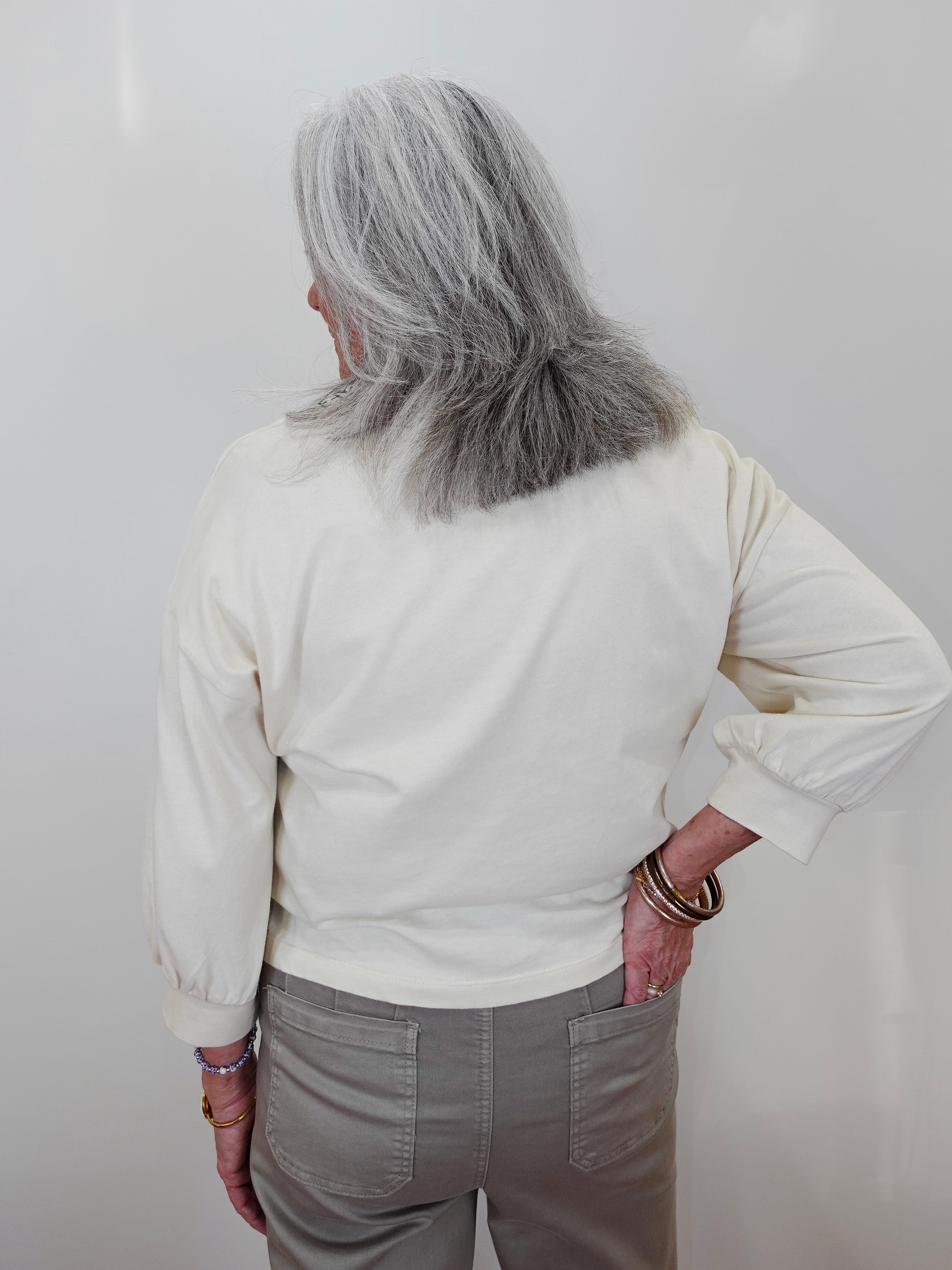 An older woman with long gray hair is shown from behind, wearing a cream-colored long-sleeve top and gray pants, standing against a plain light background.