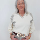 An older woman with long gray hair stands against a plain light background wearing a cream-colored long-sleeve sweater with gray floral embroidery on the shoulders and a V-neck trim, paired with gray trousers and a wide brown belt with a chunky gold buckle.