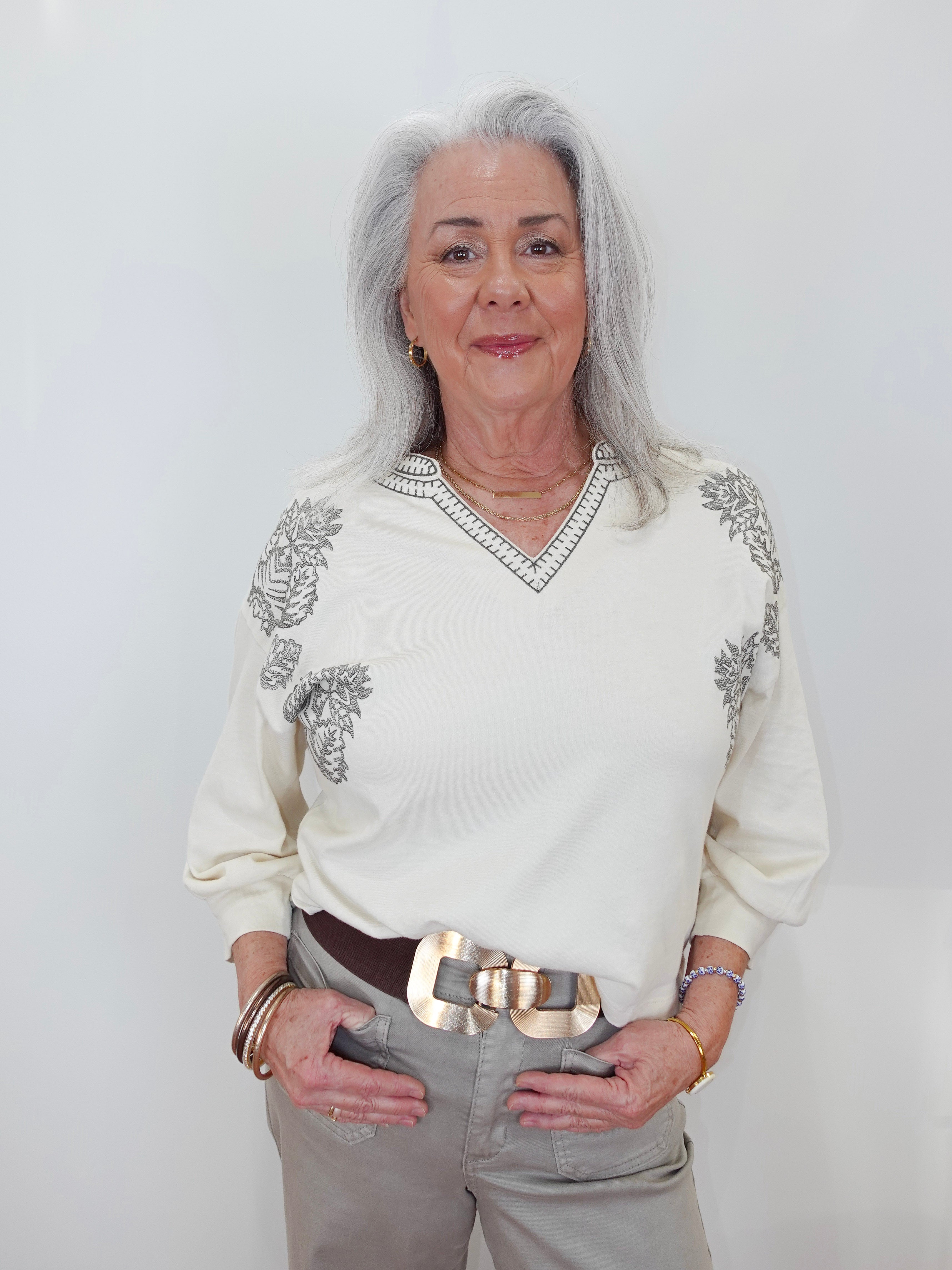 An older woman with long gray hair stands against a plain light background wearing a cream-colored long-sleeve sweater with gray floral embroidery on the shoulders and a V-neck trim, paired with gray trousers and a wide brown belt with a chunky gold buckle.
