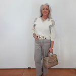 An older woman with gray hair stands against a white wall wearing a cream long-sleeve knit sweater with gray floral embroidery on the shoulders, light gray trousers, a wide gold-tone belt, and carrying a taupe handbag.