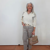 An older woman with gray hair stands against a white wall wearing a cream long-sleeve knit sweater with gray floral embroidery on the shoulders, light gray trousers, a wide gold-tone belt, and carrying a taupe handbag.