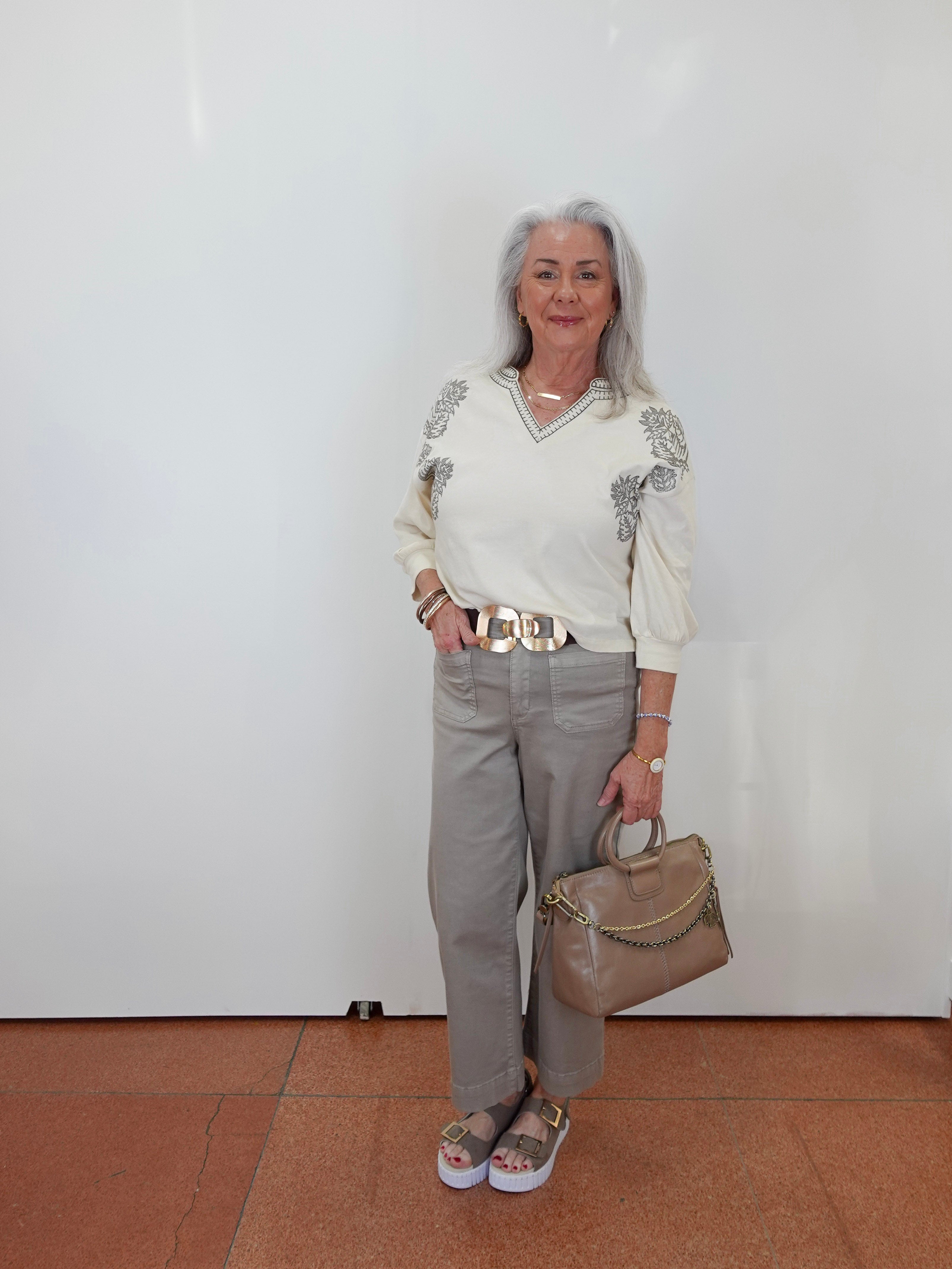 An older woman with gray hair stands against a white wall wearing a cream long-sleeve knit sweater with gray floral embroidery on the shoulders, light gray trousers, a wide gold-tone belt, and carrying a taupe handbag.