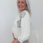 An older woman with long gray hair is shown from the waist up, wearing a cream-colored long-sleeve top with gray embroidered floral details on the shoulders and upper chest, paired with gray pants. She is smiling and looking back over her shoulder against a plain white background.