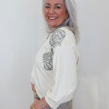 An older woman with long gray hair is shown from the waist up, wearing a cream-colored long-sleeve top with gray embroidered floral details on the shoulders and upper chest, paired with gray pants. She is smiling and looking back over her shoulder against a plain white background.