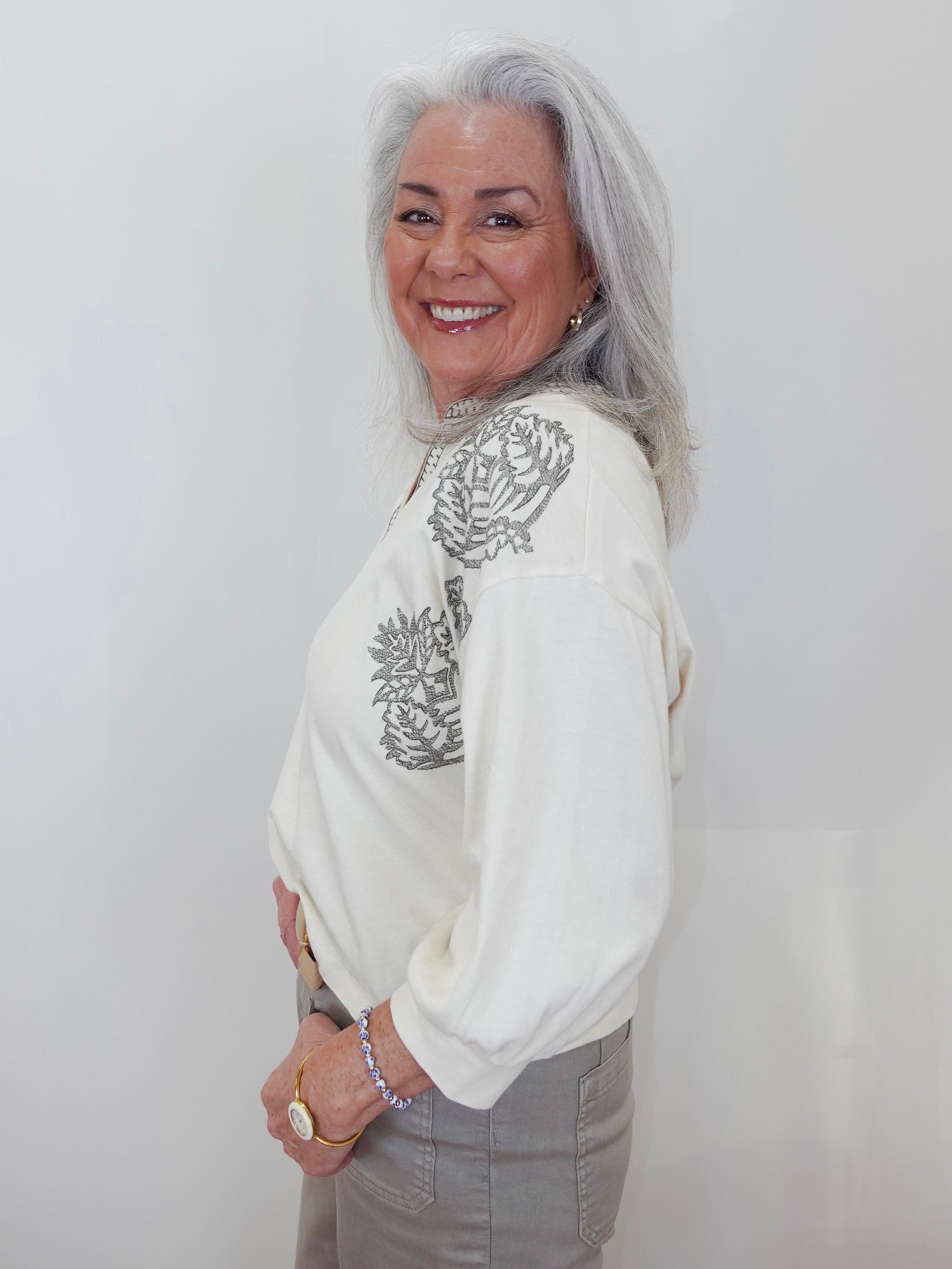 An older woman with long gray hair is shown from the waist up, wearing a cream-colored long-sleeve top with gray embroidered floral details on the shoulders and upper chest, paired with gray pants. She is smiling and looking back over her shoulder against a plain white background.