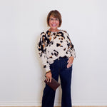 Woman wearing a patterned sweater and dark jeans against a white wall.