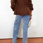 A person shown from the back wearing a brown jacket and light blue straight-leg jeans, standing against a white wall on a terracotta tiled floor.