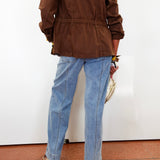 A person shown from the back wearing a brown jacket and light blue straight-leg jeans, standing against a white wall on a terracotta tiled floor.