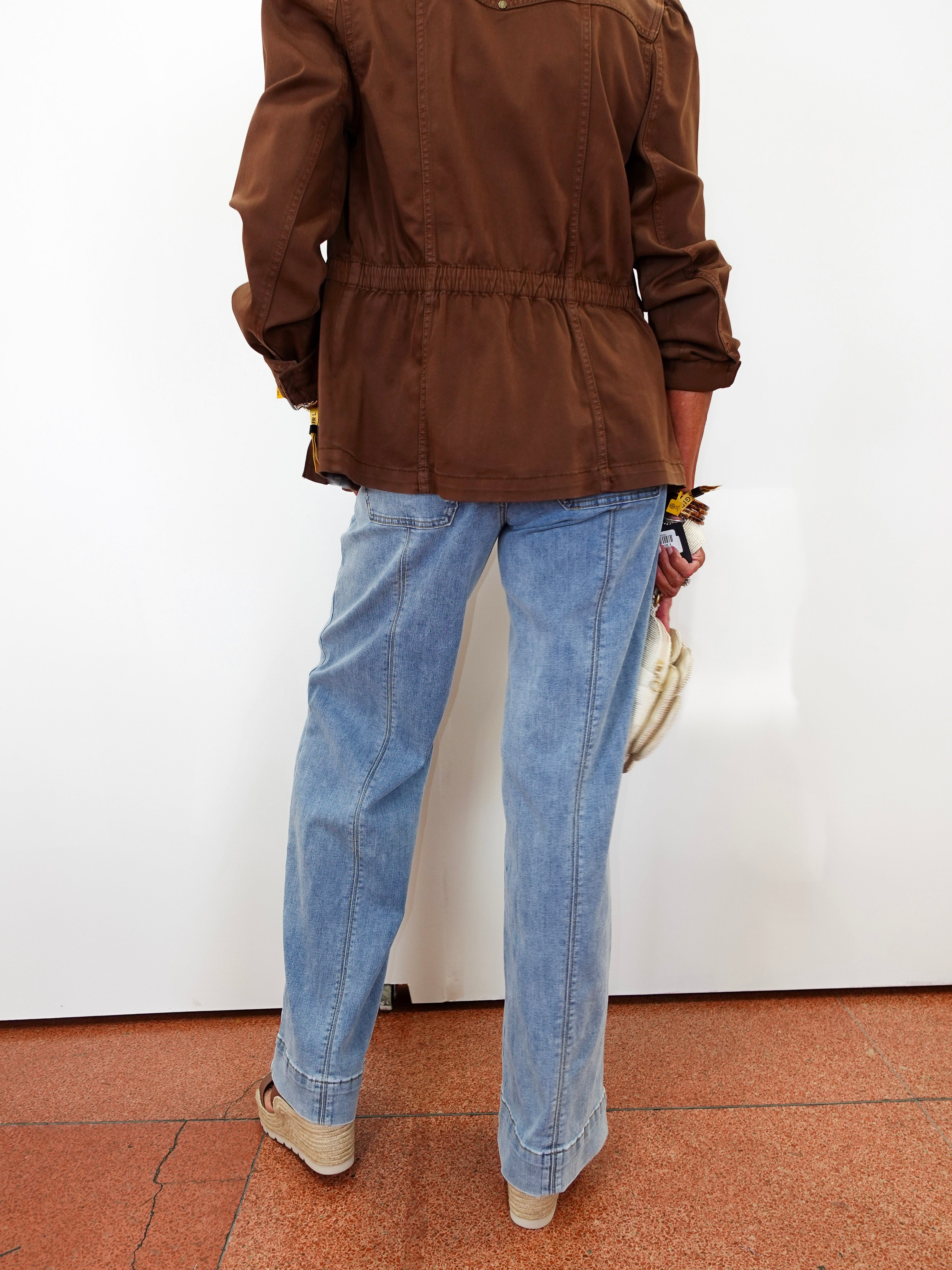 A person shown from the back wearing a brown jacket and light blue straight-leg jeans, standing against a white wall on a terracotta tiled floor.