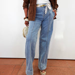 A person wearing light-wash wide-leg blue jeans with a white top, a brown jacket, and platform sandals, standing against a plain white wall.