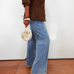 A person wearing a brown jacket, wide-leg light blue denim jeans, and beige wedge sandals, holding a cream fabric flower-shaped handbag against a plain white wall.