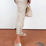 Person wearing beige pants with gold buttons, holding a beige handbag, standing on a red floor.