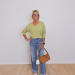Person wearing a green sweater and blue jeans, holding a brown handbag against a white wall.