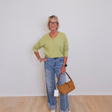 Person wearing a green sweater and blue jeans, holding a brown handbag against a white wall.