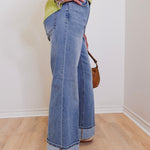 Person wearing blue jeans and a green top, standing on a wooden floor with a white wall background.