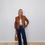 Person wearing a brown leather jacket, white top, blue jeans, and holding a brown handbag against a white wall.