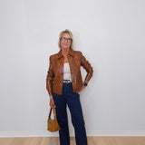 Person wearing a brown leather jacket, white top, blue jeans, and holding a brown handbag against a white wall.