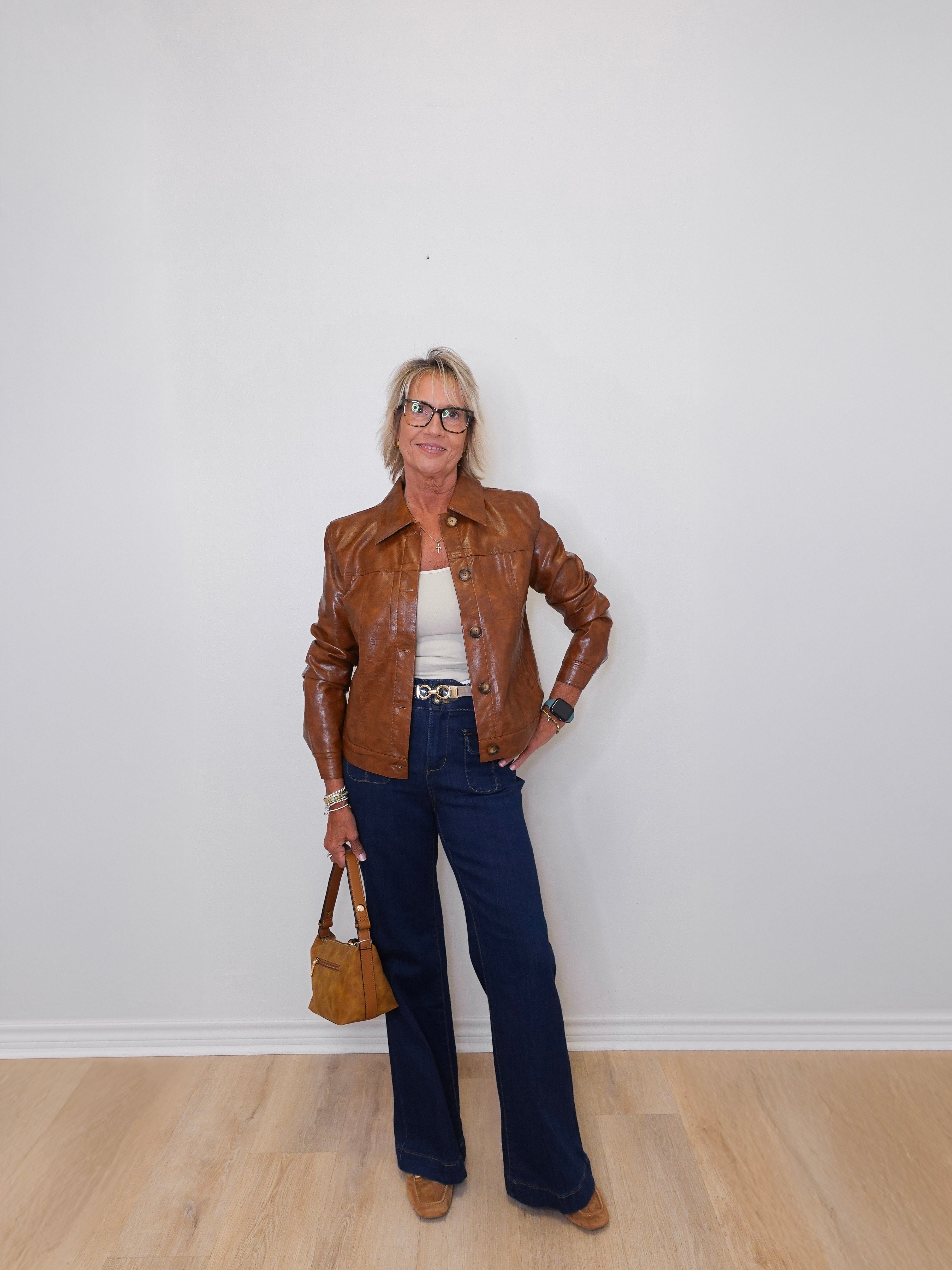 Person wearing a brown leather jacket, white top, blue jeans, and holding a brown handbag against a white wall.