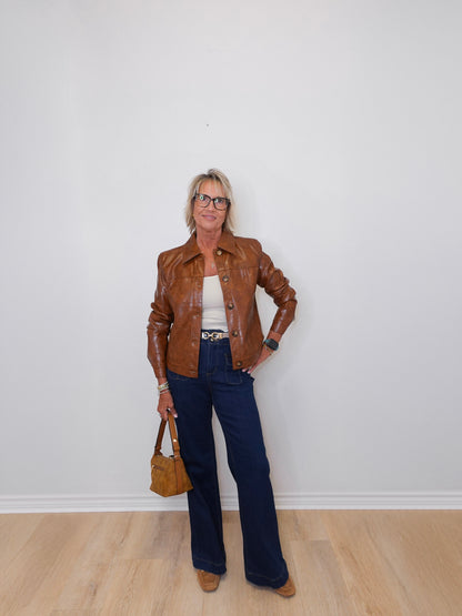 Person wearing a brown leather jacket, white top, blue jeans, and holding a brown handbag against a white wall.