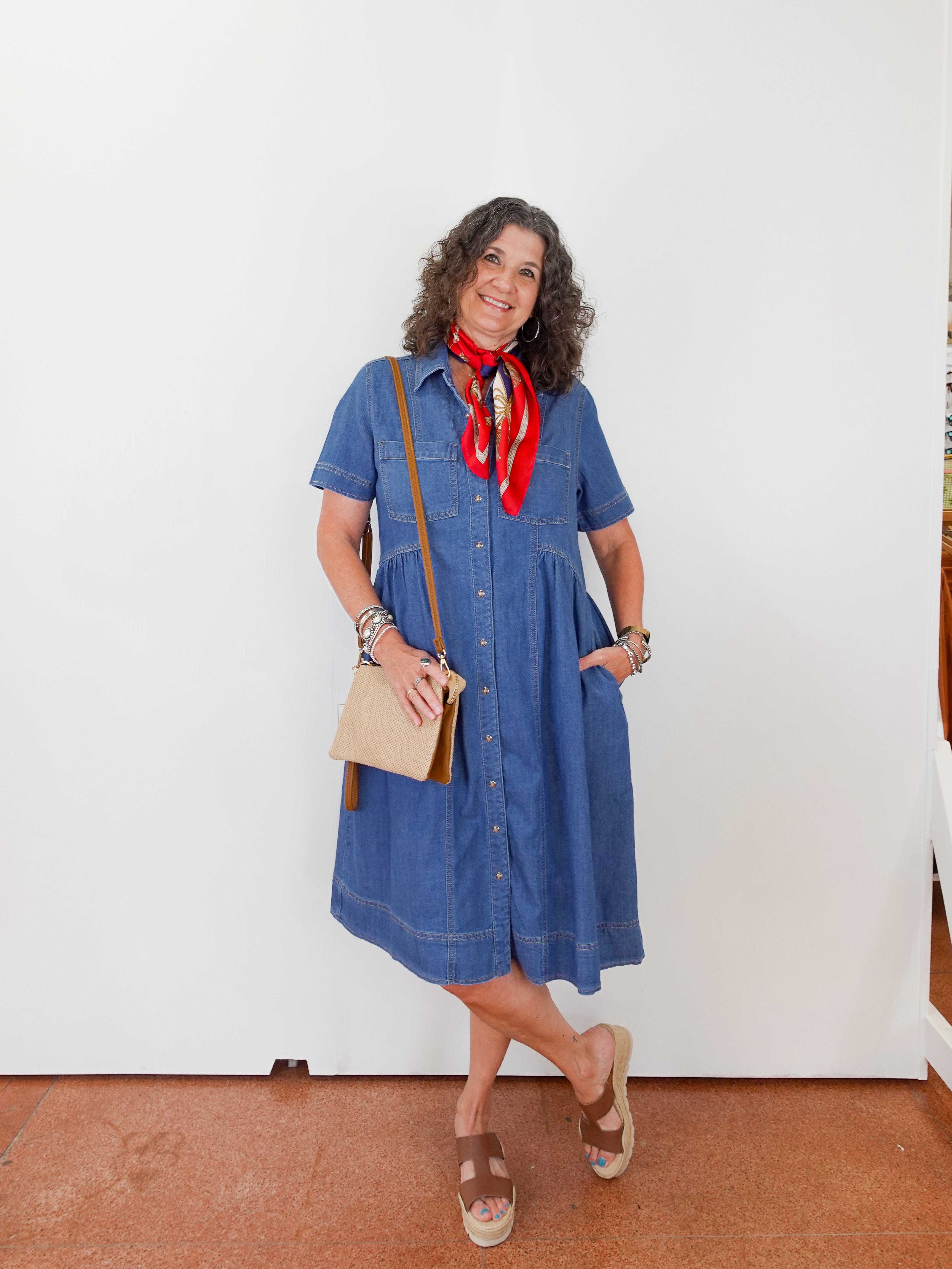 Woman wearing a blue dress with a red scarf, holding a beige bag, standing against a white wall.