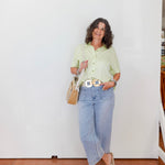 Woman wearing a light green shirt and blue jeans standing against a white wall.