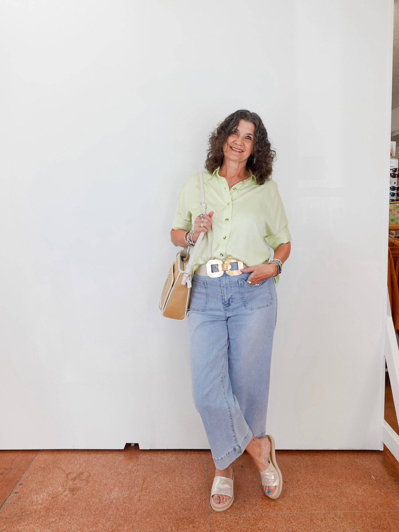 Woman wearing a light green shirt and blue jeans standing against a white wall.