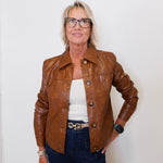 Person wearing a brown leather jacket, white top, and dark jeans against a white wall.