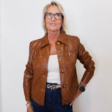 Person wearing a brown leather jacket, white top, and dark jeans against a white wall.