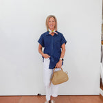 Woman in a blue shirt and white pants holding a beige handbag against a white wall.