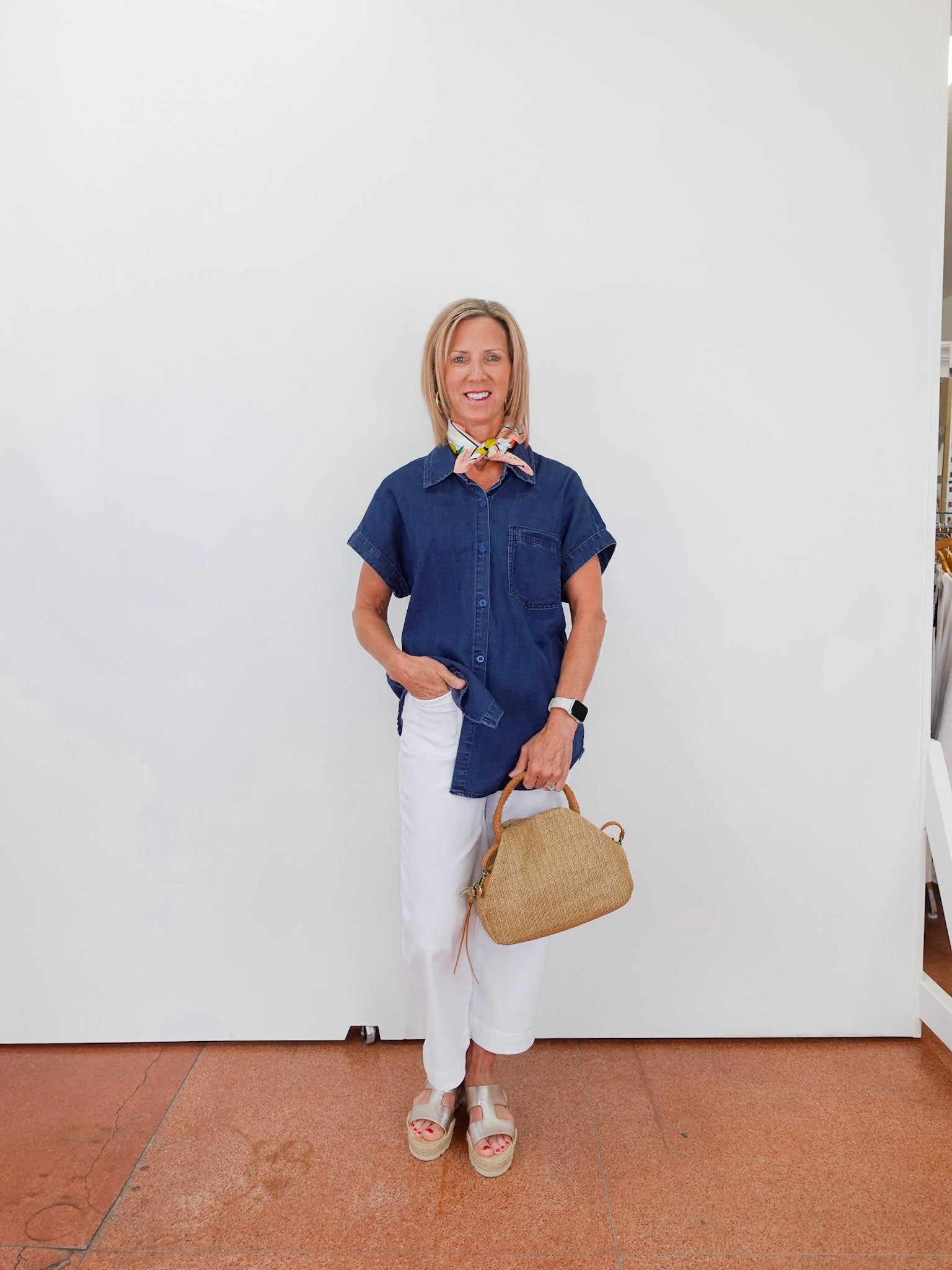 Woman in a blue shirt and white pants holding a beige handbag against a white wall.
