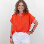 Woman wearing an orange blouse and white pants against a plain background
