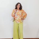 Woman wearing a colorful floral blouse and green pants against a white wall.