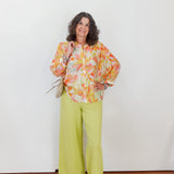 Woman wearing a colorful floral blouse and green pants against a white wall.