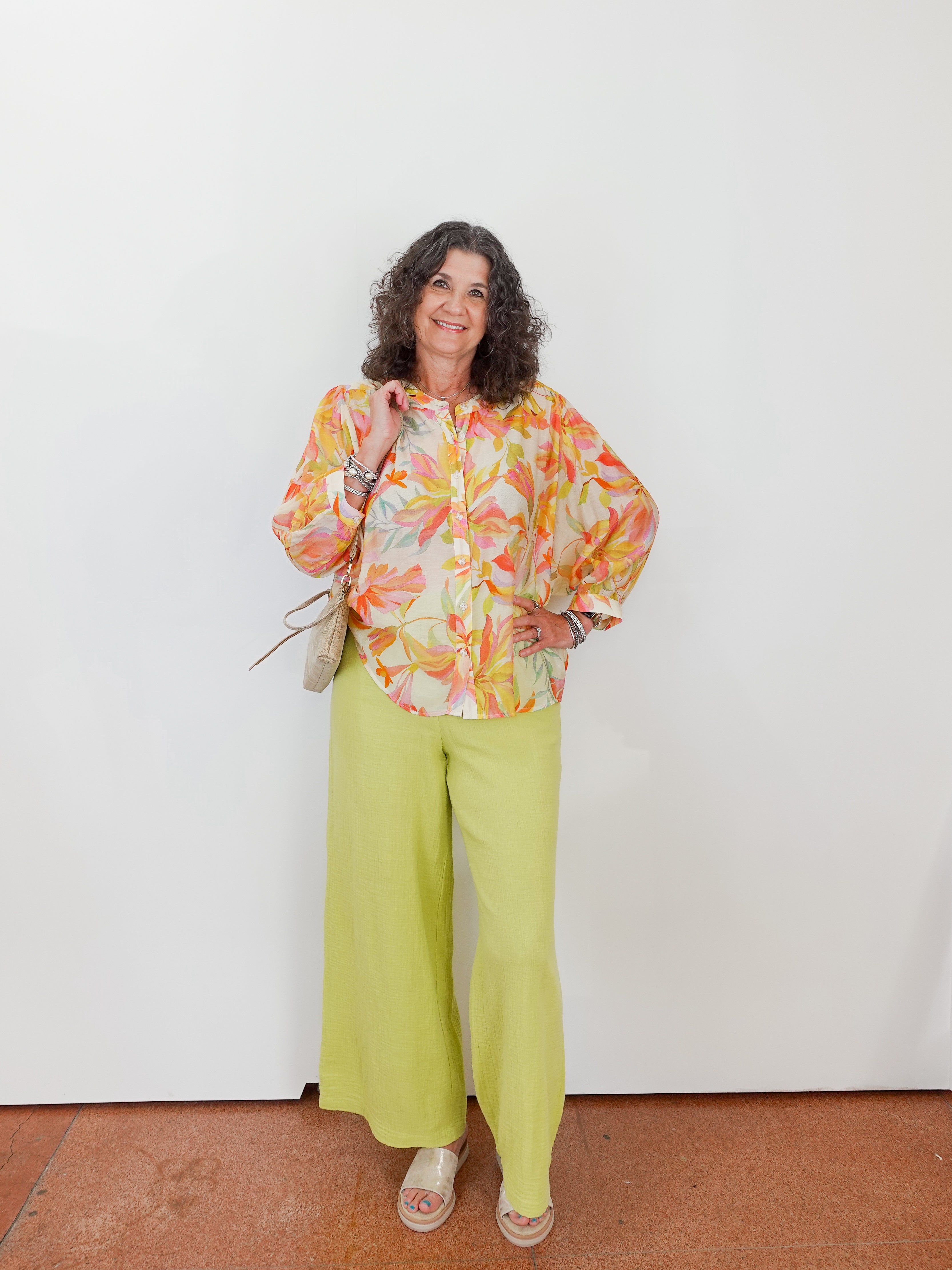 Woman wearing a colorful floral blouse and green pants against a white wall.