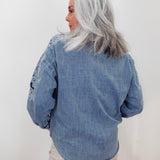 A woman with silver-gray hair is shown from behind wearing a light blue denim shirt with white embroidered details on the shoulders and sleeves, paired with beige pants, against a plain white background.
