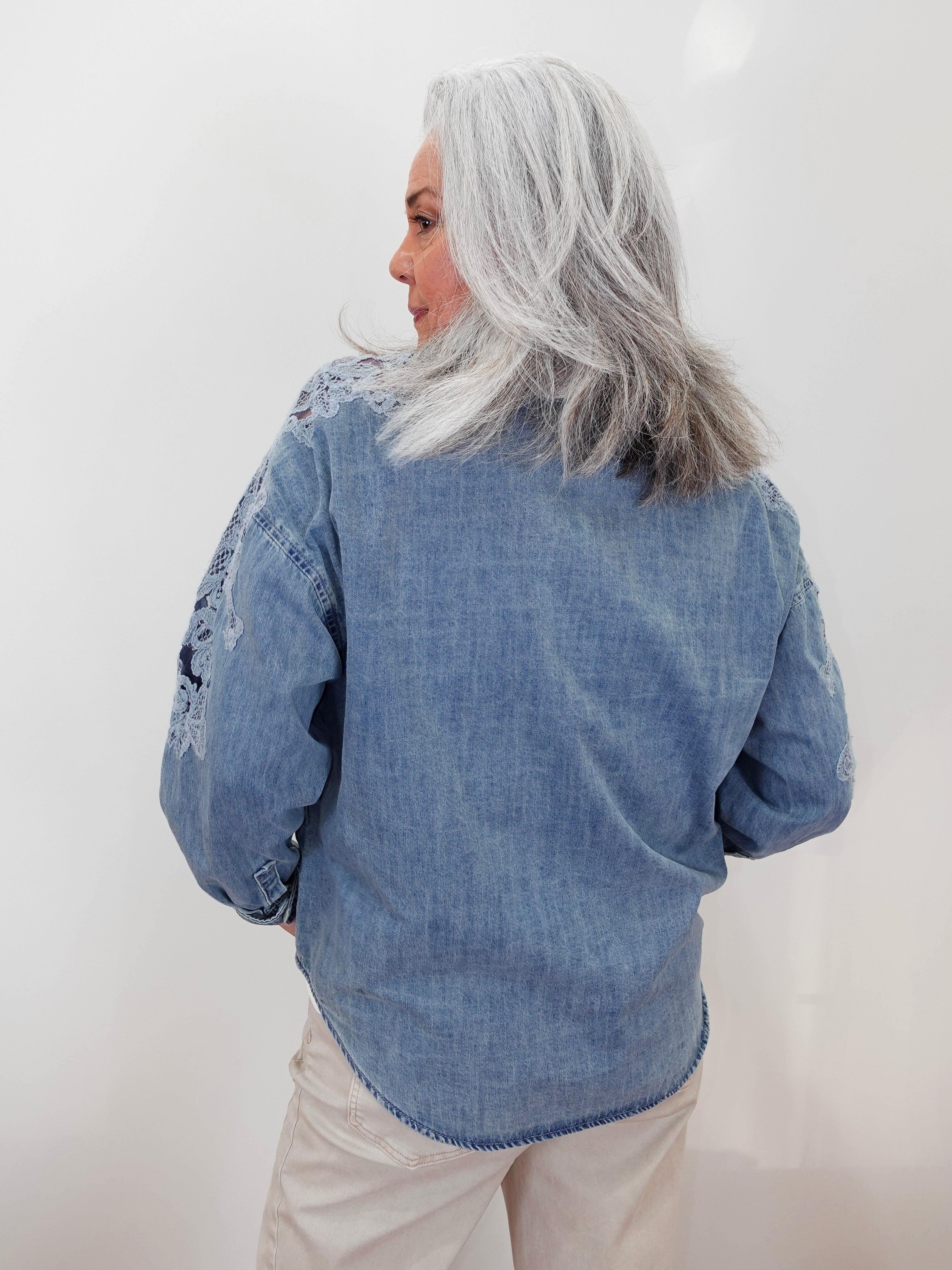 A woman with silver-gray hair is shown from behind wearing a light blue denim shirt with white embroidered details on the shoulders and sleeves, paired with beige pants, against a plain white background.