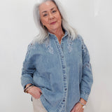 An older woman with silver hair wearing a light blue denim button-up shirt with white floral embroidery on the chest and shoulders, paired with beige pants, standing against a plain white wall.