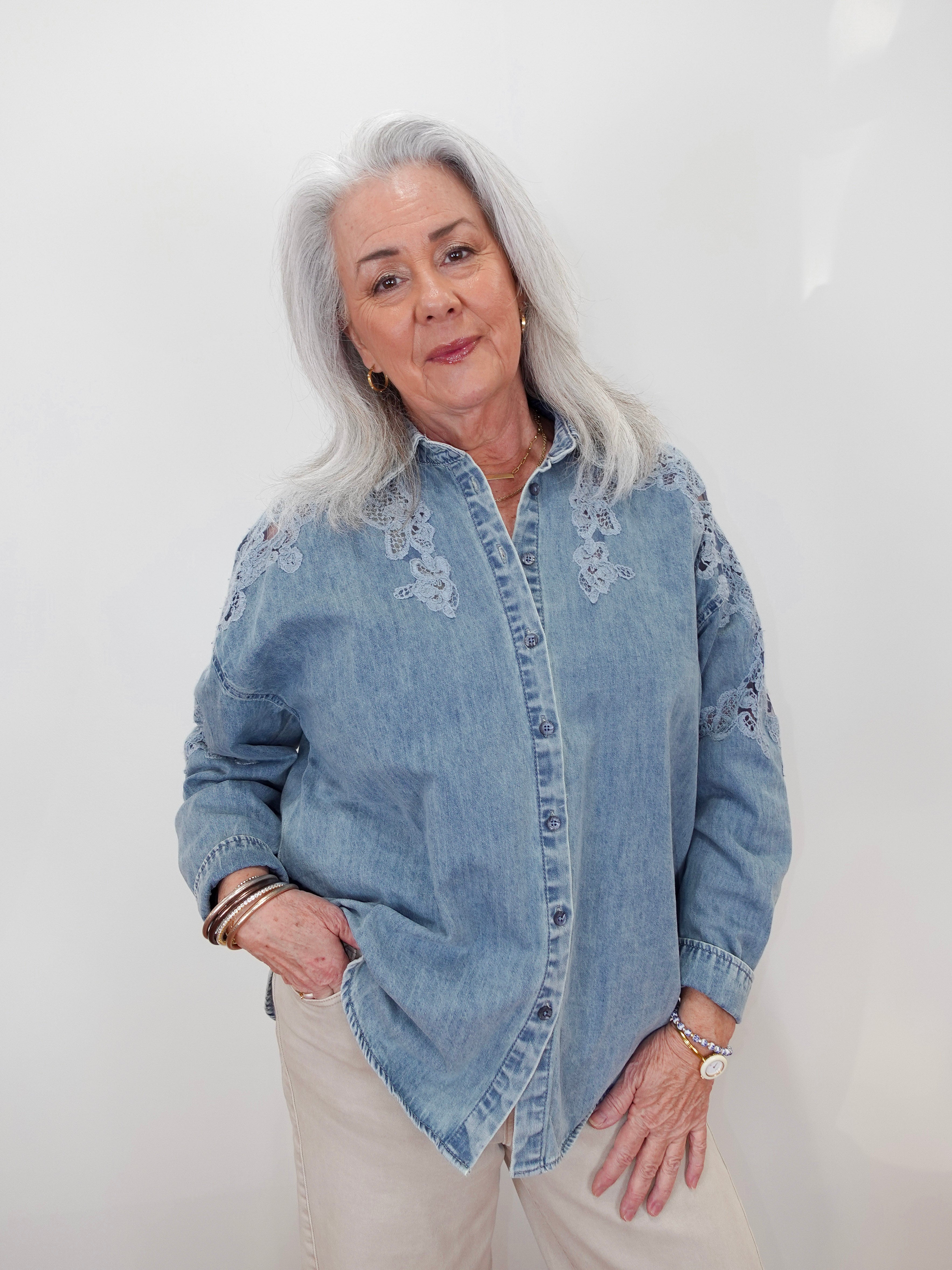 An older woman with silver hair wearing a light blue denim button-up shirt with white floral embroidery on the chest and shoulders, paired with beige pants, standing against a plain white wall.