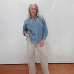 An older woman with gray hair stands against a white wall wearing a light blue denim shirt with white embroidery and beige wide-leg trousers.