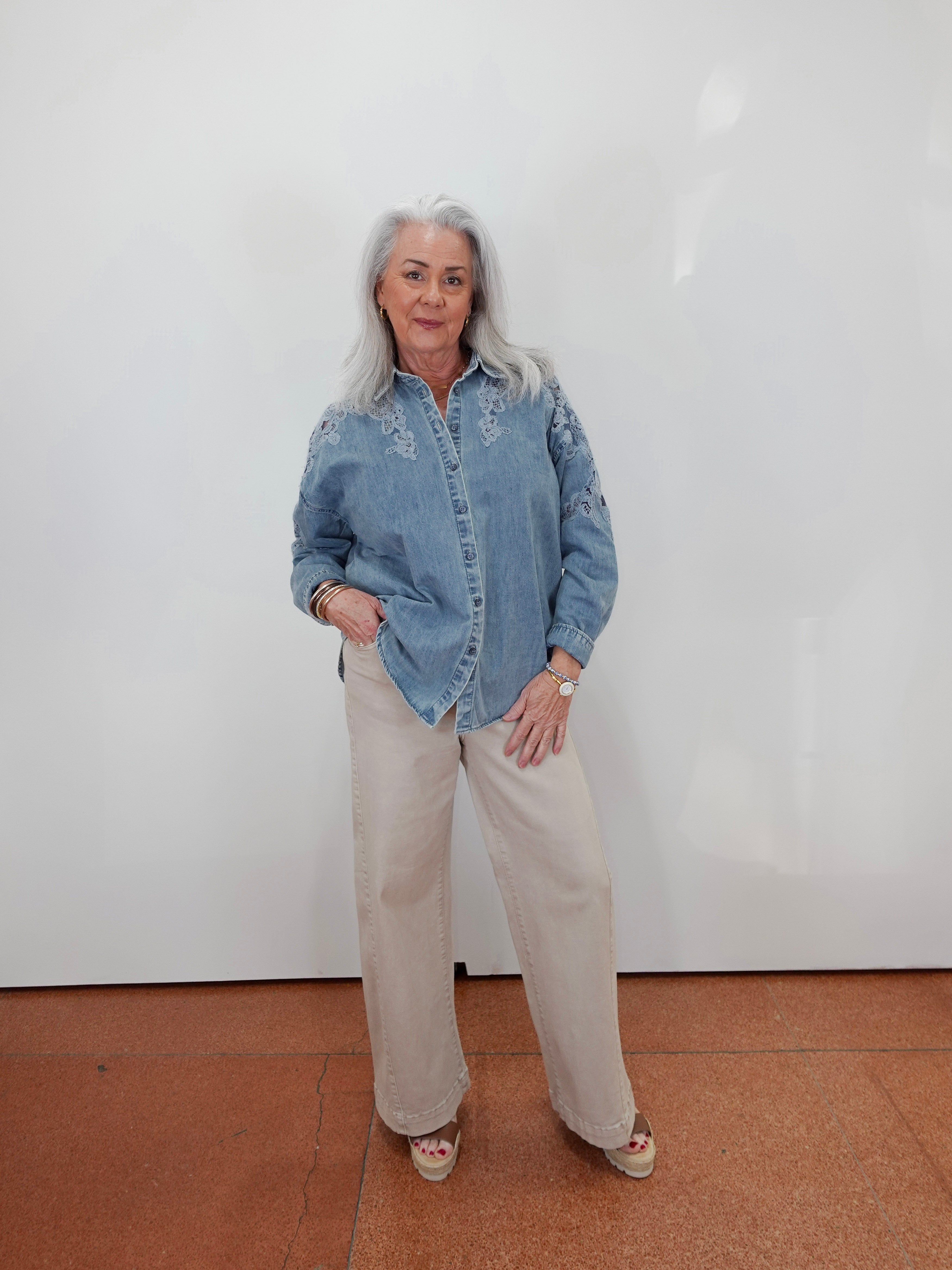 An older woman with gray hair stands against a white wall wearing a light blue denim shirt with white embroidery and beige wide-leg trousers.