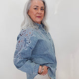 A woman with long gray hair wearing a light blue denim shirt with intricate floral lace embroidery on the sleeves, paired with cream-colored pants, standing against a plain white wall.