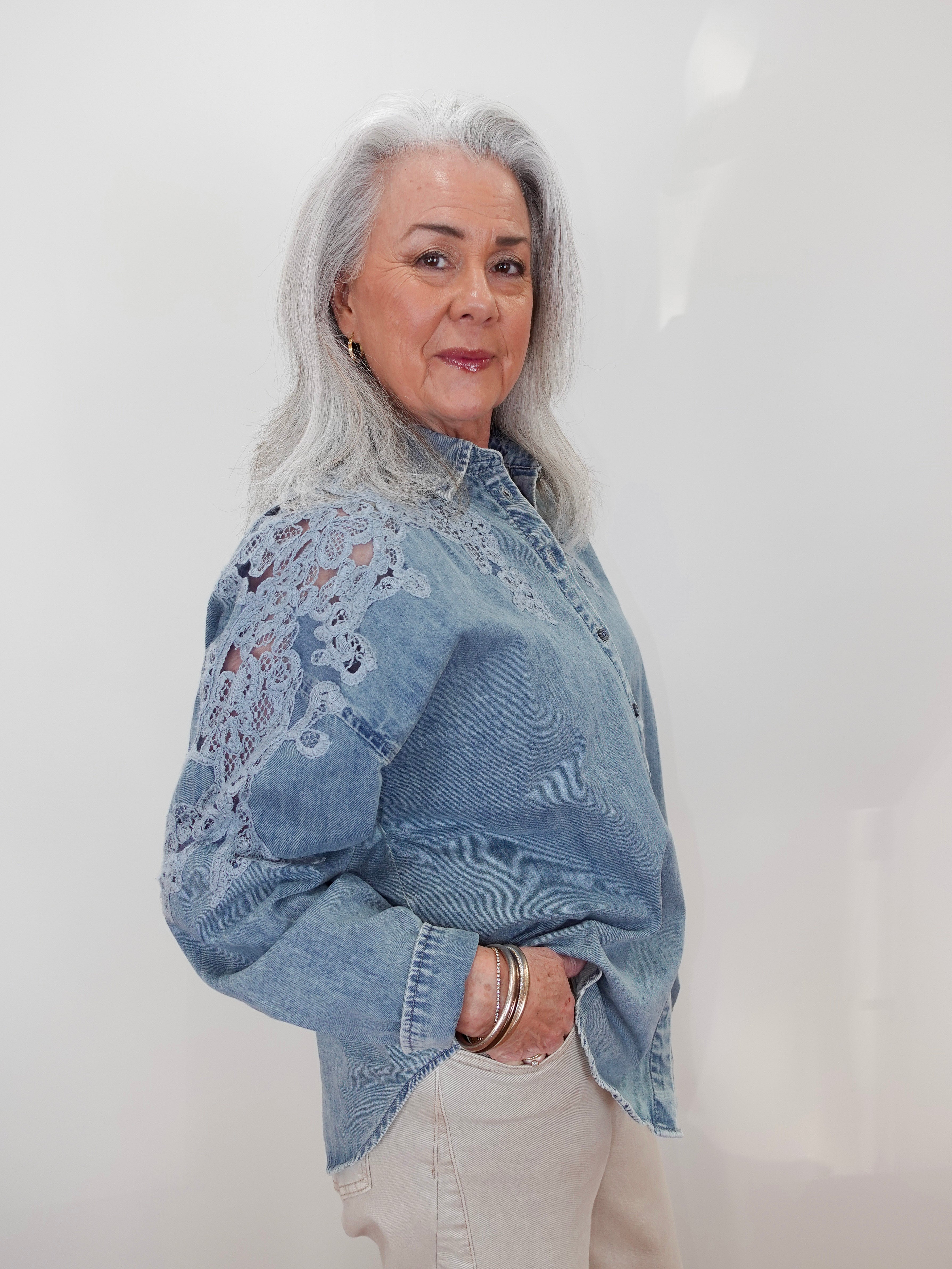 A woman with long gray hair wearing a light blue denim shirt with intricate floral lace embroidery on the sleeves, paired with cream-colored pants, standing against a plain white wall.