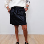 Person wearing a black leather skirt and white sweater with a graphic design, standing on a wooden floor.